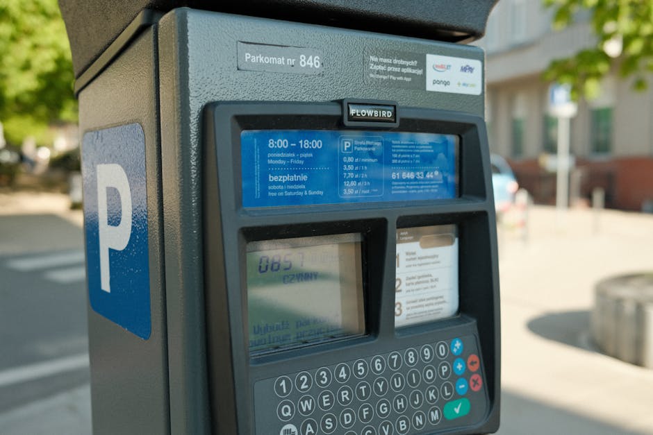 Close-up view of a parking payment machine situated on a city street, with a blue and white parking sign visible on the side. The machine displays a digital screen with parking fee details, pricing pe