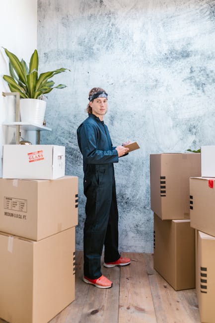 A person dressed in dark work overalls and a headband stands indoors among several cardboard boxes of various sizes, some sealed with packing tape, which are stacked on a wooden floor. The individual 