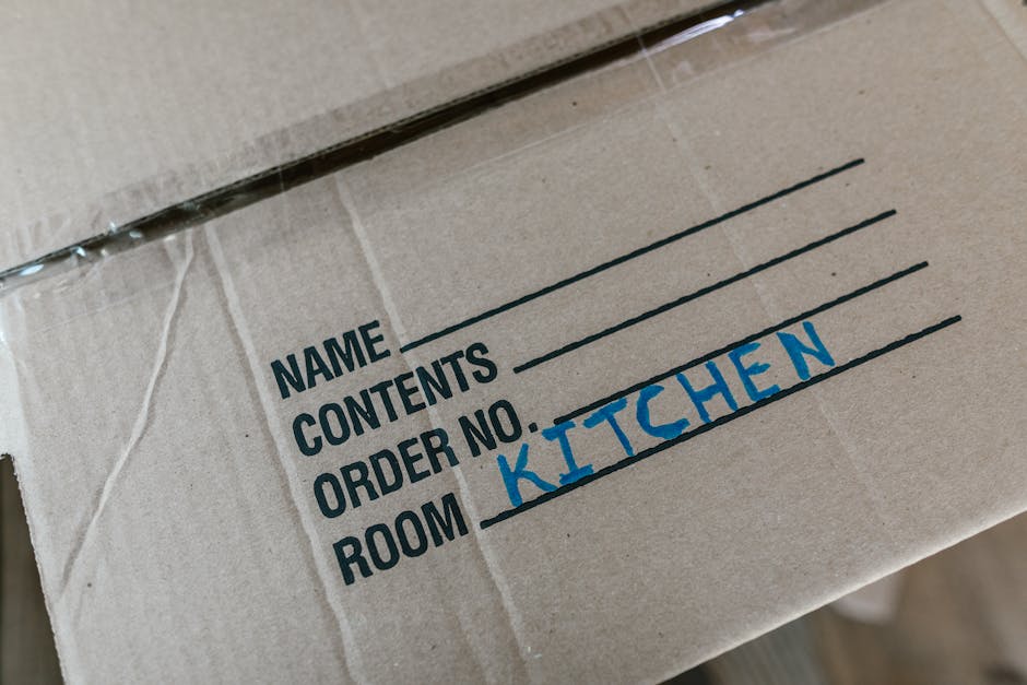 A woman with long, curly hair is standing indoors, holding two cardboard boxes stacked one on top of the other. The top box has the word 'toys' handwritten on its side. The boxes are sealed with packi