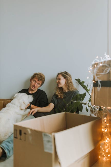 A young couple sitting on the floor of a room with light-colored walls and a large window, surrounded by open cardboard boxes and packing materials. The woman is holding a large white dog, and the man