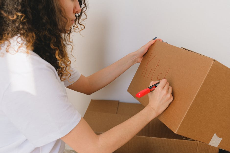 A woman with curly hair, wearing a white T-shirt, is seen writing the word 'Kitchen' on a medium-sized cardboard box with a red marker. The box is part of a stack of similar cardboard cartons, some of