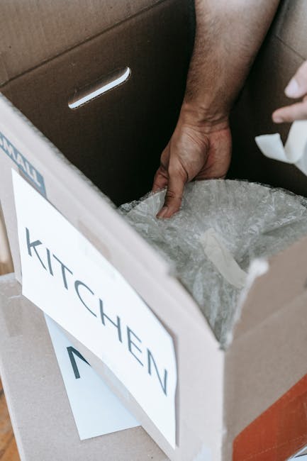 A woman with curly hair, wearing a white T-shirt, is seen writing the word 'Kitchen' on a medium-sized cardboard box with a red marker. The box is part of a stack of similar cardboard cartons, some of