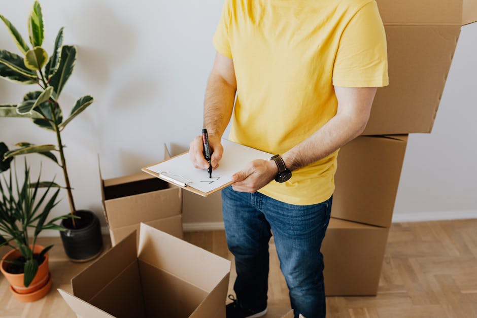 Two individuals, a man and a woman, are outside in a residential driveway during daylight, engaged in a home relocation task. The man is carrying two large cardboard boxes labeled 'BOOKS' and 'KITCHEN