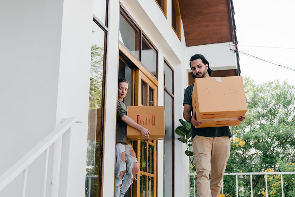 A person dressed in white trousers and an orange top is seen from behind, bending over and placing a cardboard box onto a stack of similar moving boxes inside a room with plain white walls and concret