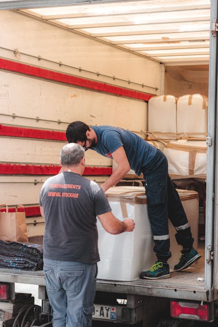 Two professional movers are inside the open back of a moving truck, engaged in a home relocation process. One mover, wearing a dark grey t-shirt with red lettering and grey pants, faces away from the