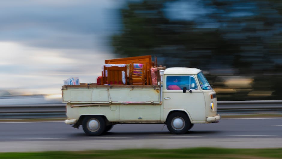Two professional movers are inside the open back of a moving truck, engaged in a home relocation process. One mover, wearing a dark grey t-shirt with red lettering and grey pants, faces away from the